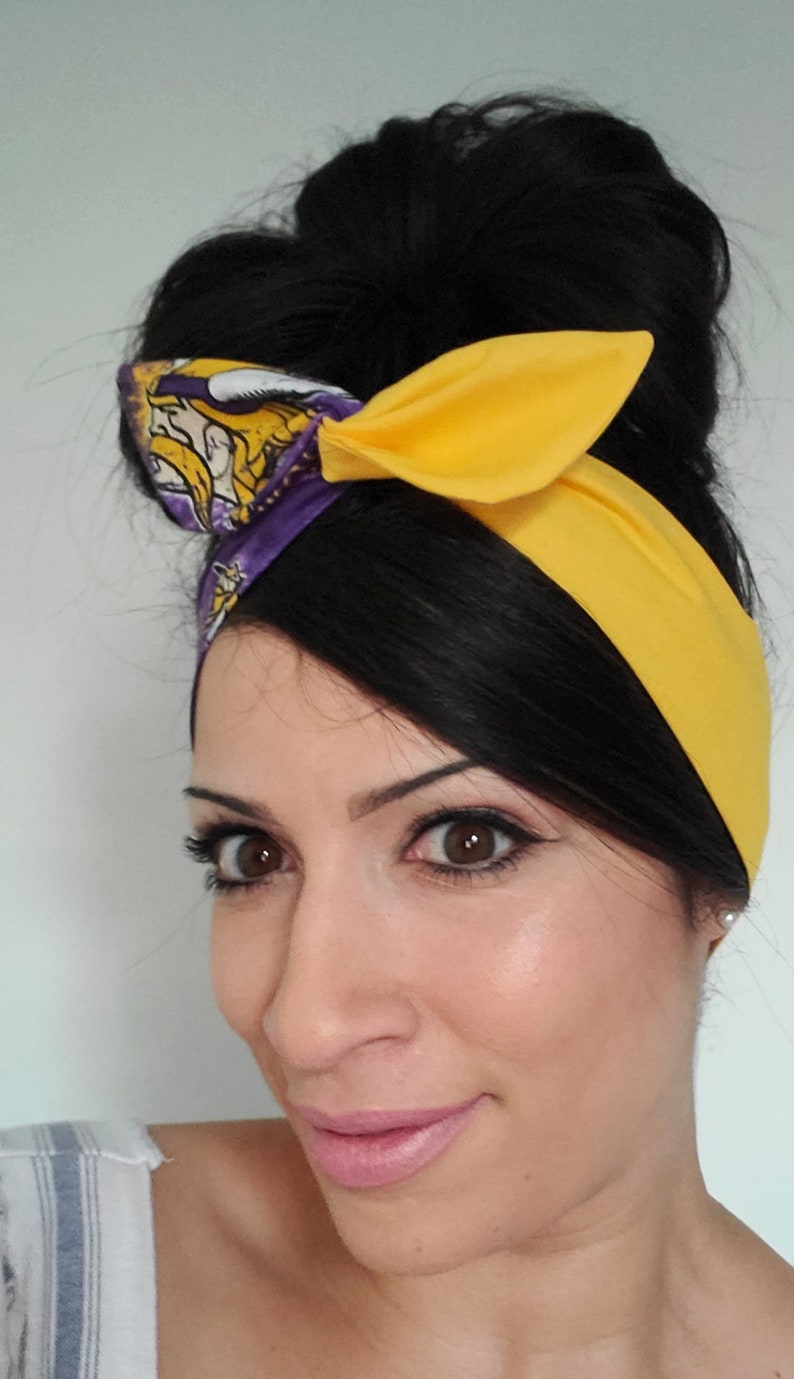 May include: A woman with dark hair styled in a bun wears a yellow headband with a purple and gold patterned section. The headband is tied in a bow at the top of her head.