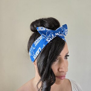 May include: A blue and white headband with the Detroit Lions logo and the words "Detroit Lions" repeated on it. The headband is tied in a bow on top of a person's head.