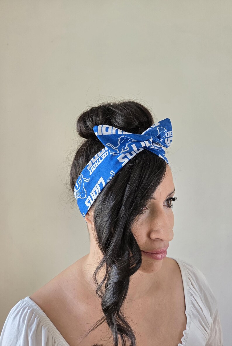 May include: A blue and white headband with the Detroit Lions logo and the words "Detroit Lions" repeated on it. The headband is tied in a bow on top of a person's head.
