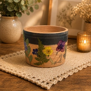 May include: A handcrafted, cylindrical planter with a natural wood finish. The planter features a dark blue band and is adorned with pressed flowers in shades of purple, yellow, and blue. It sits on a beige crocheted placemat.
