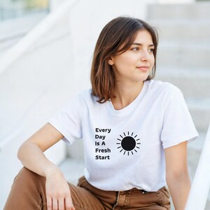 以下が含まれることがあります： 胸元に黒い太陽のグラフィックと「Every Day Is A Fresh Start」というテキストがプリントされた、白い半袖Tシャツ。クルーネックで標準的なフィット感があり、黒い文字が際立つシンプルなデザインが特徴です。