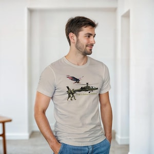 Könnte beinhalten: Ein hellbeiges Kurzarm-T-Shirt mit einem Grafikdruck eines Militärhubschraubers, zwei Soldaten in Tarnkleidung und einem Weißkopfseeadler mit Flügeln im Design der amerikanischen Flagge. Der Text "Pride in Motion in freedom" ist unter dem Hubschrauber aufgedruckt.