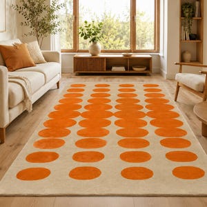 May include: A rectangular area rug featuring a repeating pattern of solid orange circles arranged in four vertical columns against a neutral beige background. The rug covers the floor space between a light-coloured sofa and a wooden armchair.