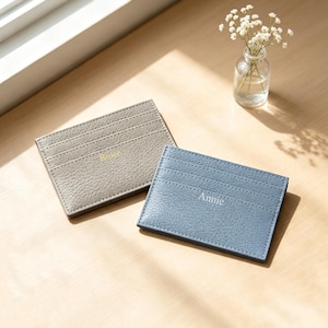 May include: Two rectangular leather card holders with a pebbled texture. One card holder is light gray with the name Rosie embossed in gold, and the other is dusty blue with the name Annie embossed in silver. Both feature multiple card slots.