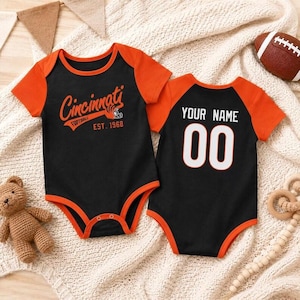 Könnte beinhalten: Zwei kurzärmelige Baby-Bodys in Schwarz und Orange. Einer zeigt den orangefarbenen Schriftzug Cincinnati Football Est. 1968. Der andere zeigt den weißen Schriftzug YOUR NAME über der Zahl 00 in Weiß mit orangefarbenem Rand. Beide haben orangefarbene Säume.