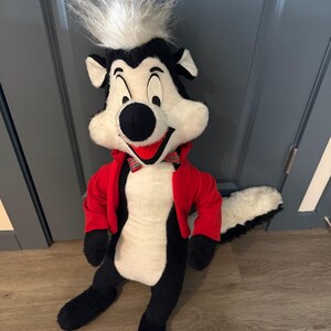 Puede incluir: Un gran peluche de un personaje de mofeta de dibujos animados. La mofeta tiene un cuerpo blanco y negro, una chaqueta roja y una pajarita a cuadros. El pelo de la mofeta es blanco y esponjoso. El juguete está sentado en un suelo de madera.