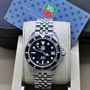 May include: A stainless steel TAG Heuer 1000 Professional wristwatch with a black dial, luminous markers, and a date window. The watch features a black rotating bezel, a metal link bracelet, and is accompanied by an international warranty card.