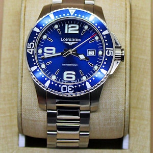 May include: A silver-tone stainless steel wristwatch with a vibrant blue dial and a matching blue rotating bezel. The watch features large white Arabic numerals at 6, 9, and 12 o'clock, a date window at 3 o'clock, and the Longines logo text.