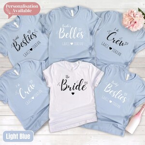 May include: A collection of five short-sleeved t-shirts in light blue and white. The shirts feature black or white printed text including phrases like The Bride, Bridal Besties, Bride's Belles, and I do Crew, along with the names Grace and Jordan.