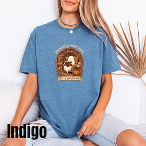 May include: A heathered indigo blue t-shirt featuring a graphic of a ghost sitting in an armchair reading a book inside a library setting. The text reads "ghosting everyone for a good book" with a pumpkin and a cup of coffee nearby.