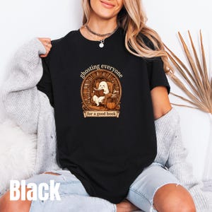 May include: A black t-shirt featuring a graphic of a ghost sitting in an armchair reading a book inside a circular library setting. The text reads "ghosting everyone" above the graphic and "for a good book" on a banner at the bottom.