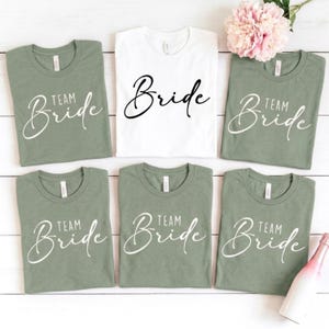 Könnte beinhalten: Fünf salbeigrüne T-Shirts und ein weißes T-Shirt in einer Gitteranordnung. Die fünf salbeigrünen Shirts zeigen den weißen Schriftzug Team Bride in Schreibschrift. Das weiße Shirt zeigt den schwarzen Schriftzug Bride in Schreibschrift.