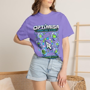 Könnte beinhalten: Ein hellviolettes T-Shirt mit einem Grafikdruck, der den Text "OPTIMISM OPERATING SYSTEM" und "CONNECTION RESTORED" enthält. Das Design zeigt einen lächelnden Cursor in der Mitte, umgeben von kreisförmigen Symbolen für Sonne, Ordner, Herz, Globus und Pflanze.