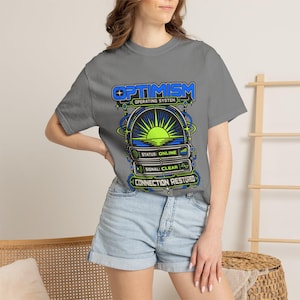May include: A grey t-shirt featuring a graphic print with the text OPTIMISM OPERATING SYSTEM, STATUS: ONLINE, SIGNAL: CLEAR, and CONNECTION RESTORED. The design includes a stylised sun rising over a horizon line, surrounded by blue and green circuitry.