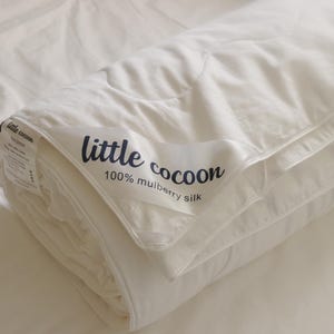 Könnte beinhalten: Eine gefaltete weiße Bettdecke mit einem Satinetikett, das den dunkelblauen Schriftzug little cocoon 100% mulberry silk trägt. Die Bettwäsche hat eine glatte, weiche Textur und ist ordentlich zusammengerollt, wobei der Markenname sichtbar ist.