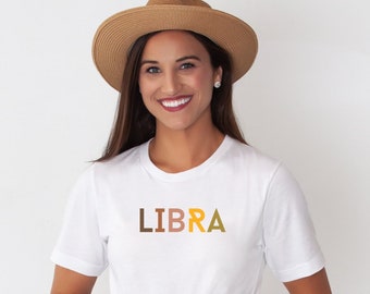 Puede incluir: Una camiseta blanca con la palabra "LIBRA" impresa en la parte delantera en letras marrones, naranjas y amarillas. La persona que lleva la camiseta sonríe y lleva un sombrero de paja.
