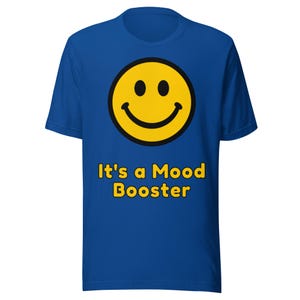 Pode incluir: Uma t-shirt de manga curta azul-real com um gráfico grande de um rosto sorridente amarelo com contorno preto centrado no peito. Abaixo do rosto sorridente, o texto "It's a Mood Booster" está impresso em letras amarelas negrito com contorno preto.