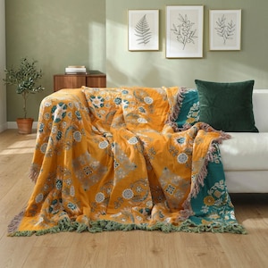 Può includere: Una coperta color giallo senape caratterizzata da un motivo floreale e botanico ripetuto nelle tonalità verde acqua, bianco e azzurro. La coperta presenta una superficie testurizzata con bordi sfrangiati lungo il fondo e i lati, su un divano bianco.