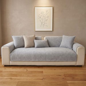 Può includere: Un divano color crema con una fodera in tessuto grigio chiaro drappeggiata sulla seduta e sullo schienale. Il divano è completato da due cuscini quadrati grigio chiaro, un cuscino quadrato beige e un cuscino rettangolare grigio chiaro al centro.