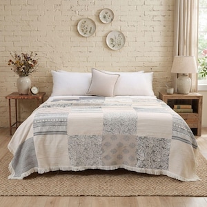 Può includere: Un copriletto patchwork caratterizzato da una varietà di motivi grigi e bianchi, inclusi disegni floreali, paisley e a righe, rifinito con un bordo sfrangiato. Il copriletto è drappeggiato su un letto con due cuscini bianchi e uno beige.