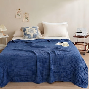 May include: A solid deep blue crinkled cotton blanket draped over a bed. The blanket features a textured, multi-layered fabric surface. It is positioned on a bed with white pillows and a decorative square pillow with a blue and white star pattern.