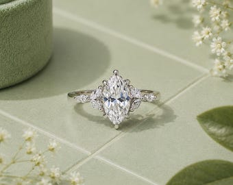 Peut inclure: Une bague argentée dotée d'une grande pierre centrale taille marquise, entourée de plus petites pierres taille marquise de chaque côté. L'anneau est fin et métallique, avec un sertissage à griffes maintenant les pierres facettées et claires.