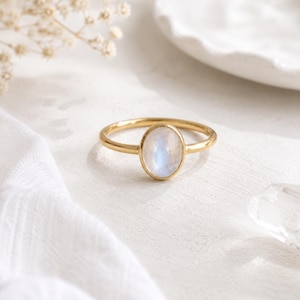 May include: A gold ring featuring a smooth, oval-shaped moonstone set in a simple metal band. The gemstone displays a soft, iridescent blue glow against the polished gold setting, creating a clean and classic appearance for this piece of jewellery.