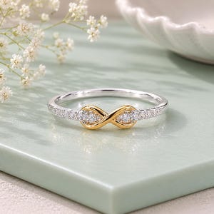 Peut inclure: Une bague bicolore avec un symbole infini central en or orné de petites pierres claires. L'anneau est en métal argenté et est agrémenté d'une rangée de minuscules pierres claires de chaque côté du motif infini central.