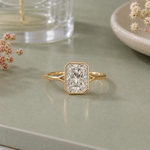 May include: A gold ring featuring a rectangular radiant-cut centre stone secured in a bezel setting. The band is thin and polished, with a split shank design that meets the central stone setting, creating a clean and structured appearance.