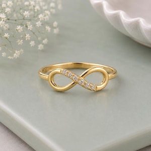 Puede incluir: Un anillo de oro con un diseño de símbolo de infinito. Un lado del bucle de infinito está adornado con cinco pequeñas piedras transparentes de corte redondo engastadas en la banda de oro pulido, creando una silueta metálica delicada.