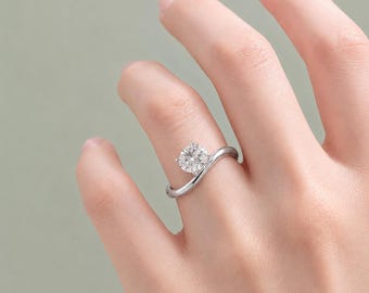 Peut inclure: Une bague argentée ornée d'une pierre précieuse ronde, transparente et à facettes, fixée par quatre griffes. L'anneau présente une finition lisse et polie avec une courbe asymétrique en forme de vague menant à la pierre centrale.