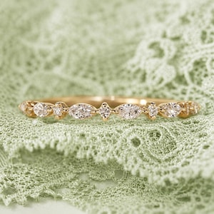 May include: A gold band ring featuring a series of alternating marquise-cut and round-cut clear stones set along the top. The metal has a polished finish, and the stones are arranged in a repeating pattern across the front of the thin band.