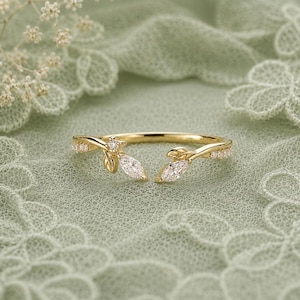 May include: A gold open-style ring featuring a branch-like design with two marquise-cut clear stones at the centre, accented by small round clear stones along the band, creating a delicate botanical appearance with a polished metallic finish.
