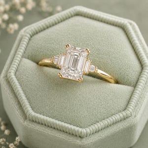 Peut inclure: Une bague en or avec une grande pierre centrale taille émeraude, flanquée de deux pierres plus petites taille baguette de chaque côté. La bague est montée sur un anneau en or fin et poli, présentant un design classique à trois pierres.