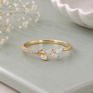 Puede incluir: Un anillo de oro con una banda delgada que presenta una hilera de pequeñas piedras redondas transparentes en un lado y un grupo de cuatro piedras transparentes de corte marquesa dispuestas en forma de hoja en el otro lado del diseño.