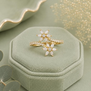 May include: A gold bypass ring featuring two flower-shaped motifs at the ends, each composed of marquise-cut clear stones surrounding a central round stone. The band is encrusted with small clear stones along the top surface of the metal.