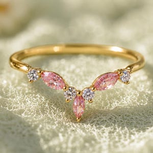 May include: A gold band ring featuring a V-shaped chevron design. The ring is set with three marquise-cut pink stones and four round-cut clear white stones arranged in a symmetrical pattern along the top of the polished gold metal band.