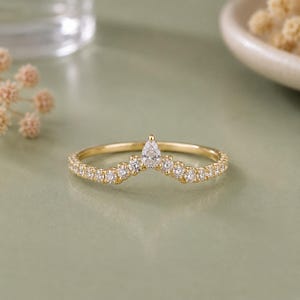 May include: A gold band ring featuring a V-shaped chevron design set with small round clear stones along the shank and a single pear-shaped clear stone at the centre point. The metal has a polished finish and the stones are set in a prong style.