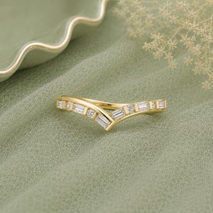 May include: A gold V-shaped chevron ring featuring a series of alternating baguette-cut and round-cut clear stones set along the band. The metal has a polished finish, and the design follows a pointed, angular silhouette that centres at the middle.