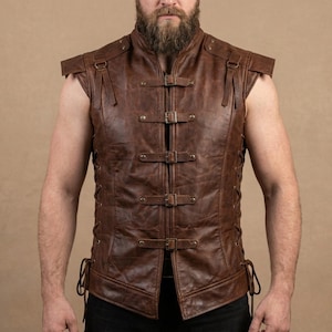 May include: A sleeveless brown leather vest featuring a high collar, four horizontal buckle straps across the front, and decorative shoulder flaps. The sides of the vest are secured with thin leather lacing that ties at the bottom hem.