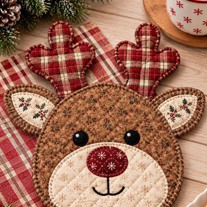 May include: A quilted fabric potholder shaped like a reindeer head. The reindeer has brown floral fabric, a cream-colored muzzle with snowflake patterns, a dark red nose, and plaid antlers. The ears are cream with small green and red holly motifs.