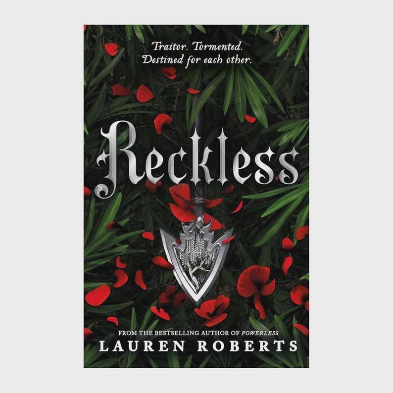 May include: The book cover for Reckless by Lauren Roberts features a silver arrowhead centred against a background of dark green foliage and scattered red petals. The title Reckless is printed in large silver lettering above the arrowhead.