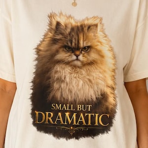 May include: An off-white short-sleeve t-shirt featuring a graphic of a fluffy, grumpy-looking cat. Below the cat, the text reads "SMALL BUT DRAMATIC" in a gold-colored serif font. The shirt has a crew neckline and a relaxed, boxy fit.