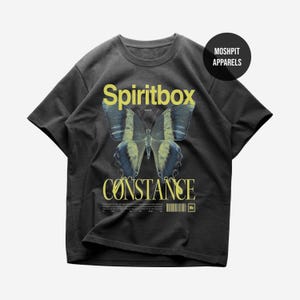 Puede incluir: Camiseta gris oscuro con un diseño de mariposa. El diseño incluye la palabra "Spiritbox" encima de la mariposa y "Constance" debajo, en amarillo. La camiseta también tiene un código de barras y el texto "Moshpit Apparels".