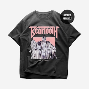 Puede incluir: Una camiseta gris carbón de manga corta con un estampado gráfico que muestra el texto BEARTOOTH en letras rosas sobre una figura metálica cromada sentada en un trono. Debajo de la figura, el texto THE SURFACE aparece en letras blancas.