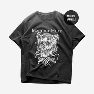 Puede incluir: Una camiseta negra de manga corta con un estampado gráfico blanco de una calavera rodeada de cuernos y serpientes. El texto "MACHINE HEAD" está impreso sobre la calavera, y un logotipo en forma de diamante con las letras "MH" está debajo. "MOSH PIT APPARELS" está en un círculo.