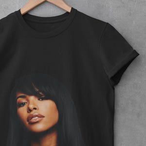 May include: A black short-sleeved t-shirt featuring a large, centered photographic print of a person with long dark hair, bangs, and a silver necklace. The garment has a classic crew neckline and is displayed on a wooden hanger.