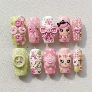 May include: A set of ten press-on nails featuring pink, green, and white designs. Decorations include 3D buttons, bows, hearts, a cartoon face, a cat face, and text reading Sweet Cute and A to Z. Patterns include gingham, polka dots, and lace.