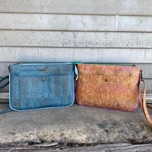 May include: Two cork crossbody bags, one blue and one pink, with a brown cork pattern. Both bags have a small metal button closure.