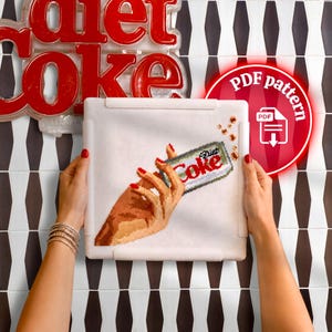 Könnte beinhalten: Ein Kreuzstich-Stickprojekt auf weißem Stoff, das in einem weißen Kunststoffrahmen gehalten wird. Das Design zeigt eine Hand mit rot lackierten Fingernägeln, die eine silberne Diet Coke Dose hält, mit kleinen braunen Blasen oben rechts.